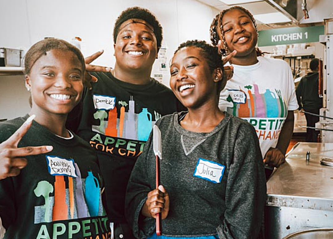 Four people looking toward the camera smiling while standing in Appetite for Change's commercial kitchen space during a Community Cooks event. Three people display peace signs and one holds a spatula.